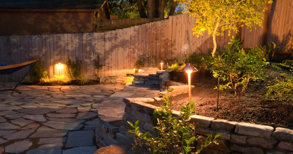 Warmly lit backyard patio with stone pavers, tiered retaining walls, landscaped garden beds, and pathway lighting illuminating trees and plants at night.