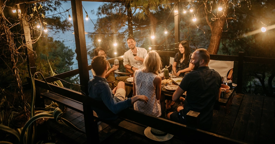 a group of friends gathered on someone’s outdoor deck with lights hanging around the perimeter