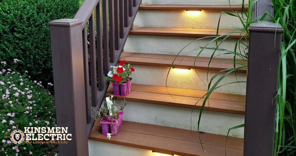 Multi-level outdoor deck with dark brown railings and softly lit stairways overlooking a neatly maintained backyard with lush landscaping and a curved concrete path.