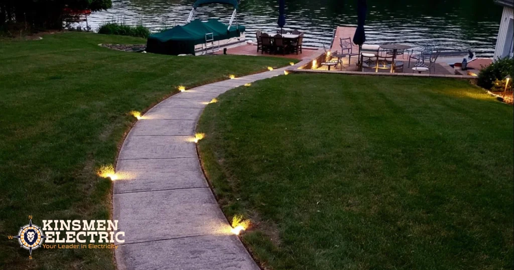 Curved concrete walkway lined with warm landscape lighting leading down a grassy lakeside yard to a dock and patio seating area beside a calm lake at dusk.
