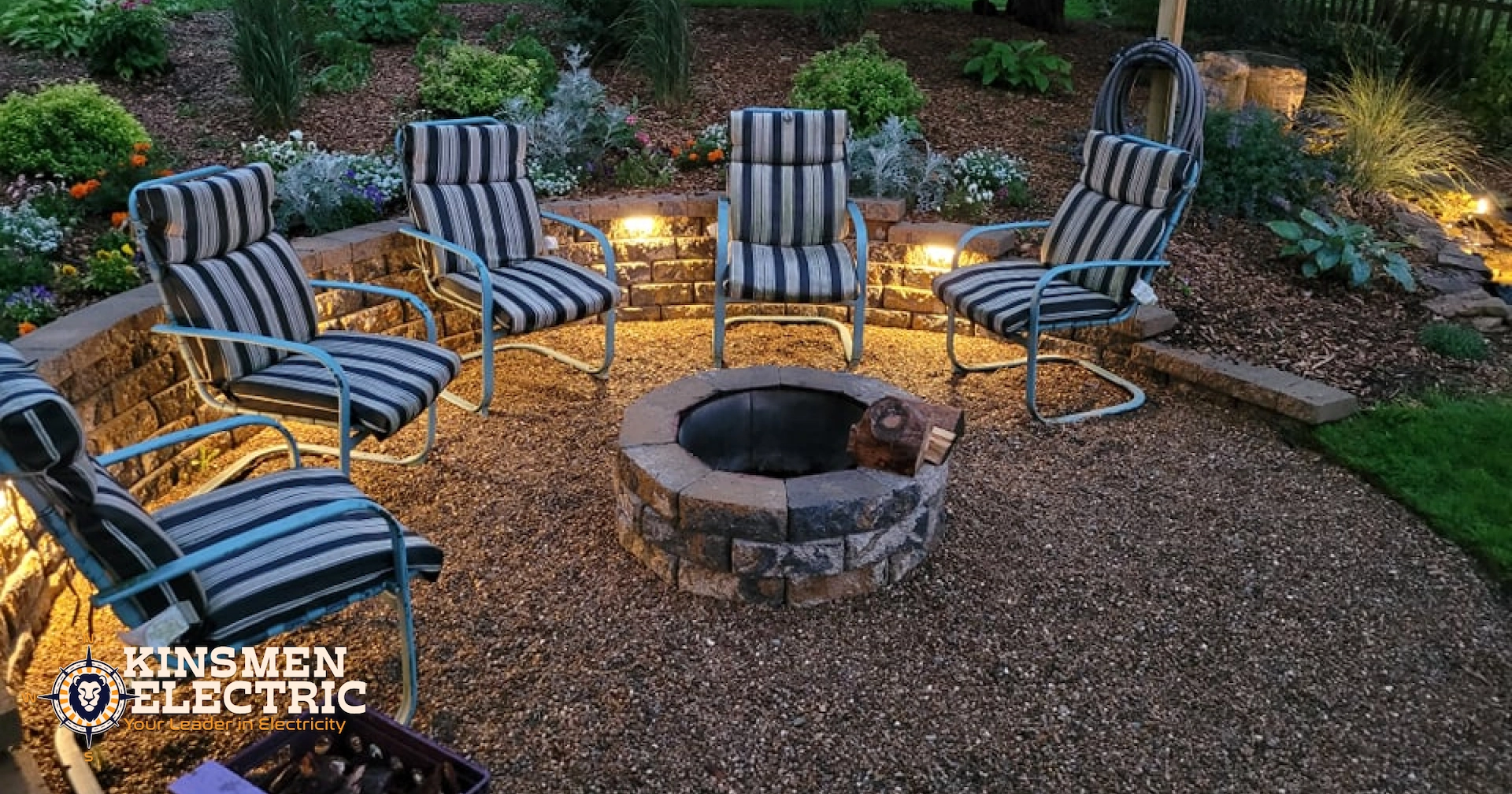 Outdoor patio with stone fire pit surrounded by striped cushion chairs on gravel, with landscape lighting and garden beds at dusk.