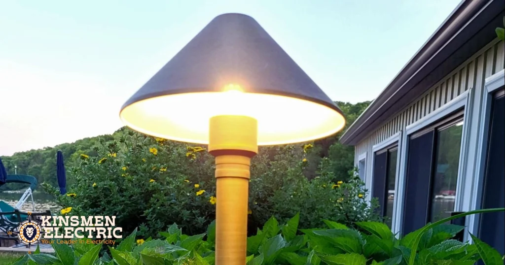 Brass landscape path light with cone-shaped shade glowing at dusk in a garden bed, with lush green shrubs and a lakeside home in the background.