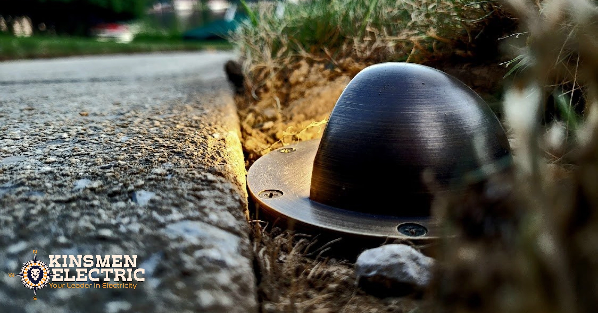 Close-up of a bronze dome-shaped in-ground well light installed along a driveway edge, casting a warm glow into ornamental grass.