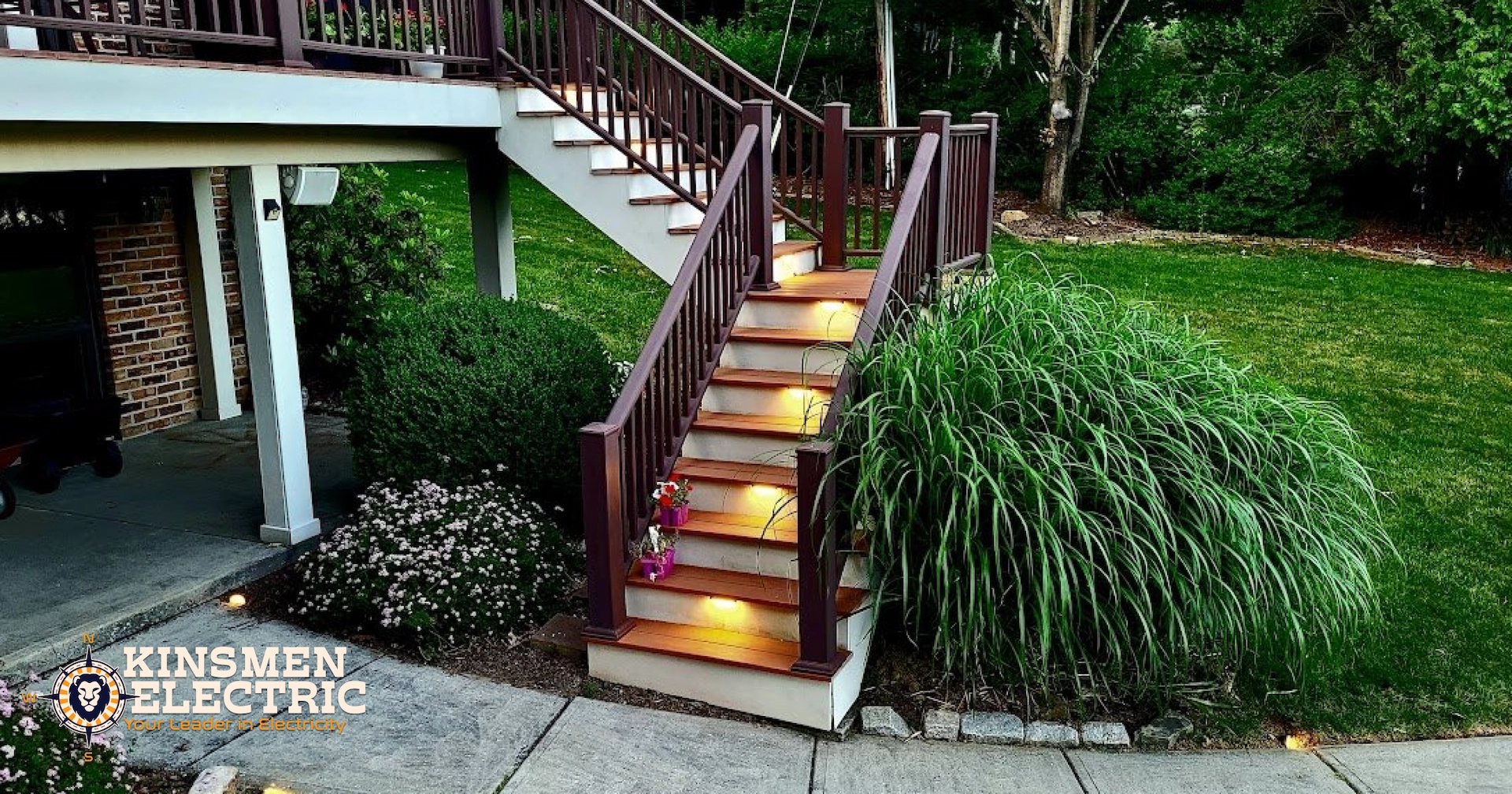 Elevated backyard deck with brown railing and illuminated wooden stairs leading to a landscaped yard with ornamental grasses, trees, and a curved concrete walkway at dusk.