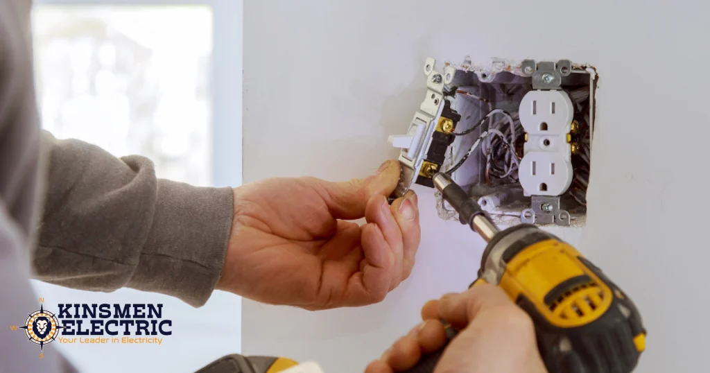 A close-up of an electrician installing a white light switch into an open electrical wall box while using a cordless drill. A newly installed white duplex outlet is visible beside the switch, with exposed wiring inside the wall.